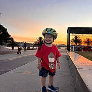 Sahas is registered to the contest to win money with this photo: child, toddler, skateboard, helmet, red_shirt, shorts, shoes, sunset, palm_trees, waterfront, sidewalk, outdoor, smiling, happy, people, urban, recreation, park, evening, play