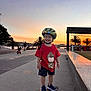 child, toddler, skateboard, helmet, red_shirt, shorts, shoes, sunset, palm_trees, waterfront, sidewalk, outdoor, smiling, happy, people, urban, recreation, park, evening, play