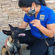 Shadow joined the competition — help win amazing prizes! woman, dog, puppy, mask, blue_shirt, sidewalk, stone_wall, leash, collar, black_hair, crouching, pet, outdoor, interaction, smiling_dog, casual_clothing, hand_gesture, bag, happy, training