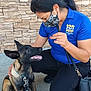 bag, black_hair, blue_shirt, casual_clothing, collar, crouching, dog, hand_gesture, happy, interaction, leash, mask, outdoor, pet, puppy, sidewalk, smiling_dog, stone_wall, training, woman