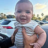 Gunner is registered to the contest to win money with this photo: baby, child, person, smile, happy, outdoor, daylight, sky, cloud, car, parking_lot, hand, striped_clothing, short_sleeve, palm_tree, holding, portrait, cute, infant, wristwatch