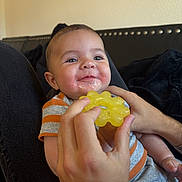Gunner is registered to the contest to win money with this photo: baby, child, teether, hand, smile, drool, striped_shirt, indoor, person, face, infant, happy, couch, black_fabric, skin, head, holding, yellow, closeup, comfort