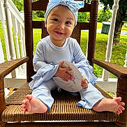 Mariposa is registered to the contest to win money with this photo: baby, child, smiling, blue_headband, bow, striped_clothing, wooden_chair, porch, outdoor, greenery, milestone_card, happy, feet, hands, face, sitting, cute, infant, natural_light, wood