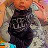 Legend joined the competition — help win amazing prizes! baby, infant, child, clothing, blanket, person, face, sitting, indoors, cute, cozy, black_clothing, gray_clothing, hand, patterned_blanket, soft_toys, portrait, young_child, warm_lighting, curious_expression
