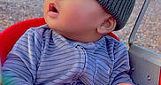 Legend is registered to the contest to win money with this photo: baby, child, swing, red_swing, hat, knit_hat, onesie, striped_clothing, hand, park, outdoor, grass, trees, curious, wide_eyes, seated, infant, playground, nature, daylight