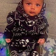 Legend is registered to the contest to win money with this photo: baby, child, infant, sitting, black_clothing, hoodie, sports_pattern, soccer_ball, basketball_hoop, blanket, indoors, curious_expression, face, feet, socks, couch, cozy, patterned_blanket, young_child, portrait