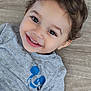 child, toddler, smiling, face, eyes, hair, grey_shirt, mickey_mouse, blue_logo, wooden_floor, happy, portrait, cute, baby, person, casual_clothing, indoors, skin, expression, young_child