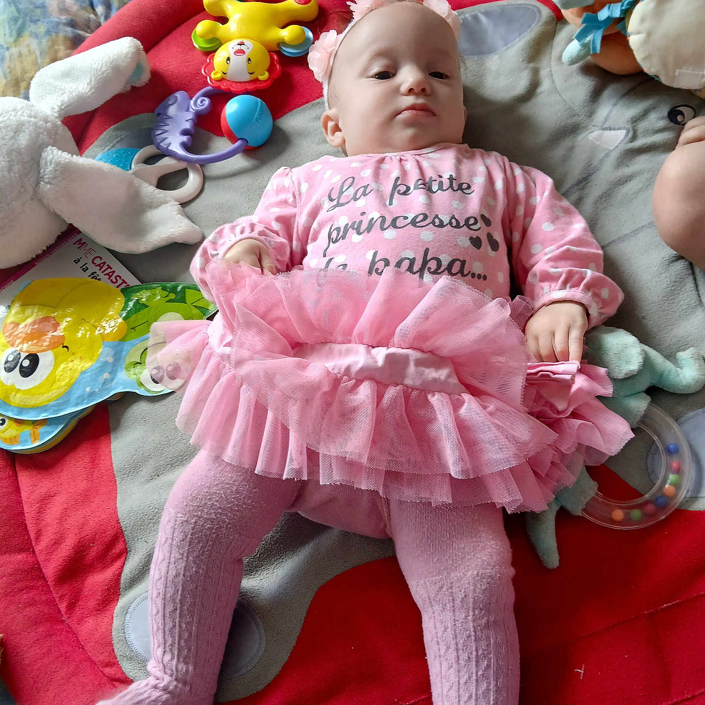 Joy participe au concours pour gagner de l'argent avec cette photo : baby, book, child, clothing, colorful, cute, floor, headband, indoor, infant, laying_down, pink_tutu, play_mat, portrait, serious_expression, soft_toys, stuffed_animal, tights, toys, young_child