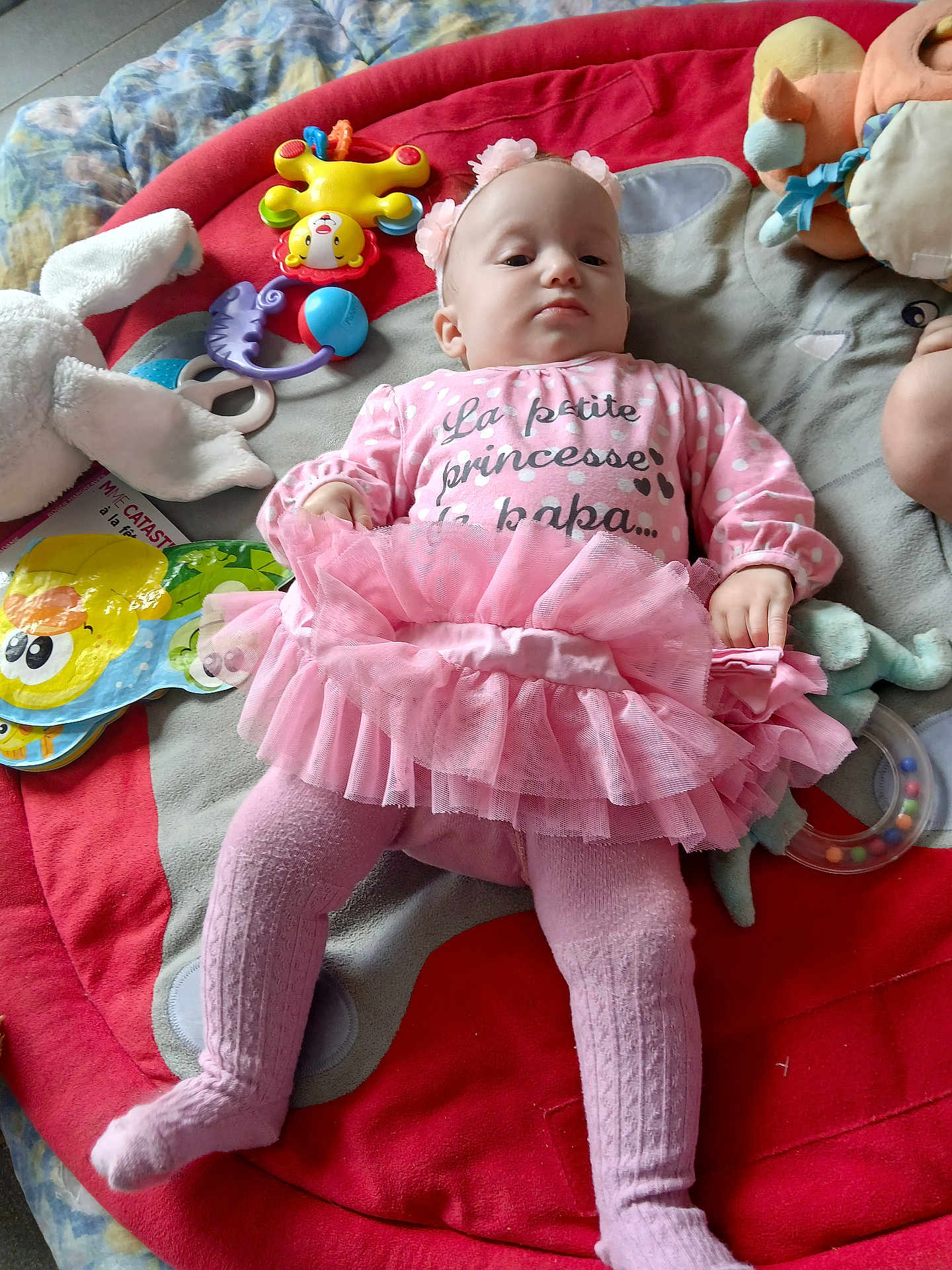 Joy participe au concours pour gagner de l'argent avec cette photo : baby, pink_tutu, tights, headband, play_mat, toys, stuffed_animal, book, indoor, child, infant, clothing, laying_down, cute, serious_expression, colorful, soft_toys, floor, young_child, portrait