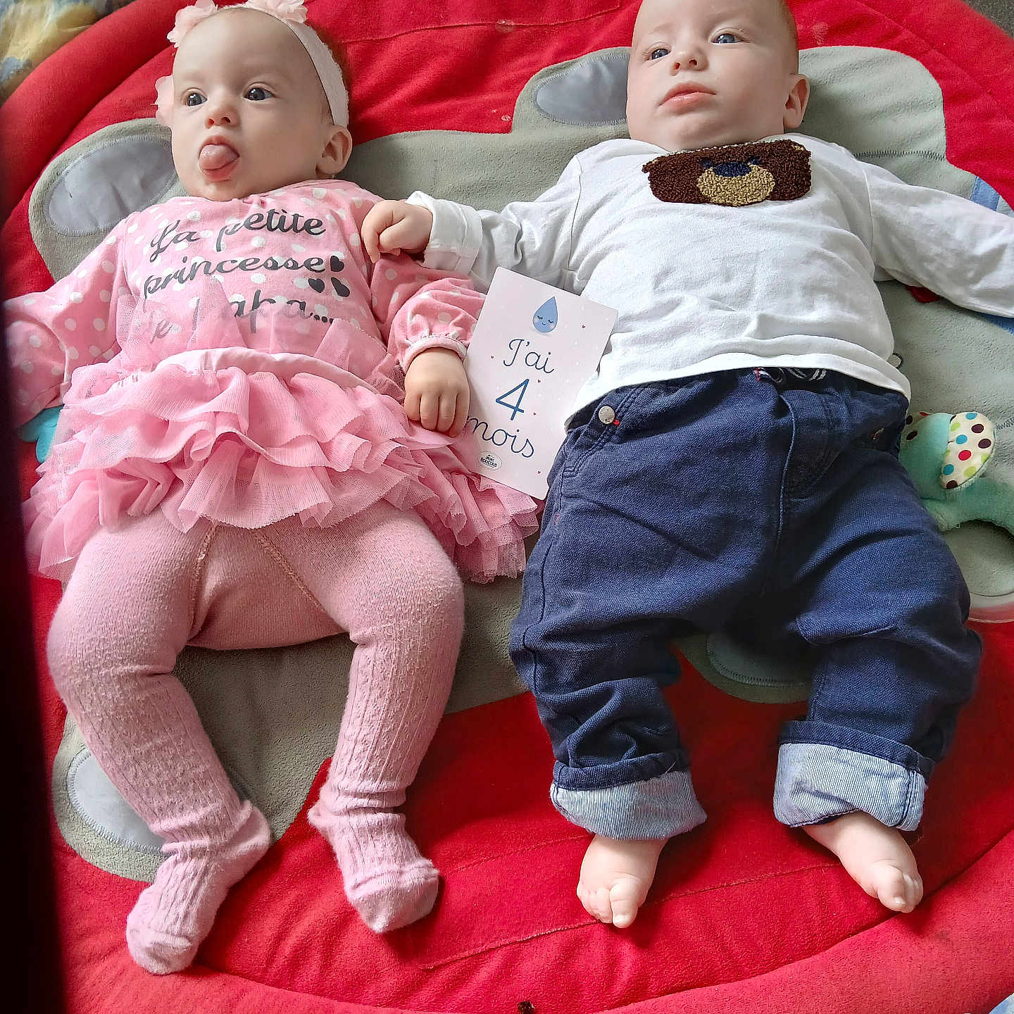 Thimothe participe au concours pour gagner de l'argent avec cette photo : baby, blanket, blue_clothing, child, cute, face, feet, floor, headband, holding_card, indoors, infant, lying_down, milestone_card, pink_clothing, playful, plush_mat, soft_toys, toddler, tongue_out