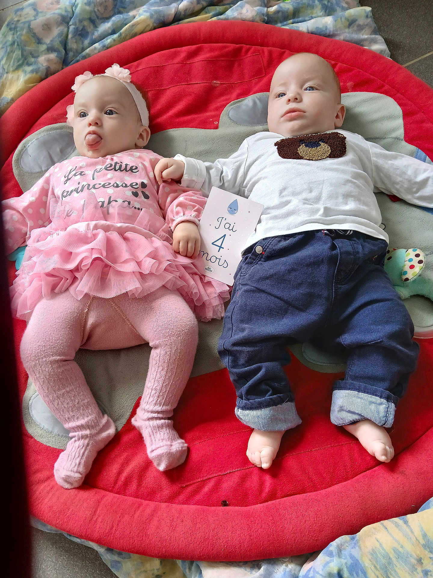 Thimothe participe au concours pour gagner de l'argent avec cette photo : baby, infant, child, pink_clothing, blue_clothing, headband, plush_mat, playful, tongue_out, holding_card, milestone_card, lying_down, cute, indoors, soft_toys, toddler, face, feet, floor, blanket