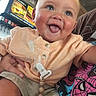 baby, child, infant, smiling, tongue_out, portrait, indoor, restaurant, seated, onesie, shorts, pacifier_clip, teething_beads, colorful_clothing, close_up, face, happy, playful, background_sign, photography