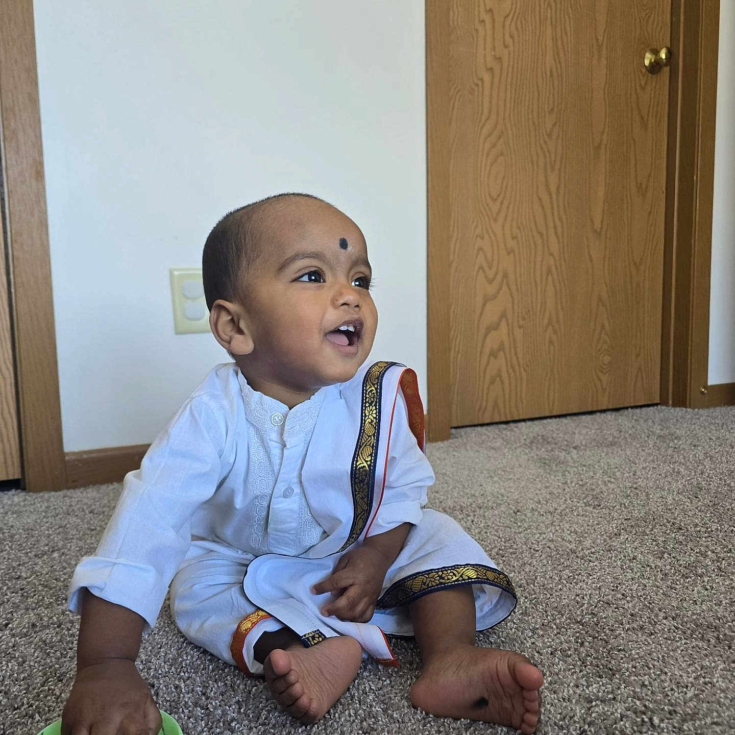 Harshavardhan joined the competition — help win amazing prizes! toddler, child, indoor, carpet, door, wall, white_clothing, traditional_clothing, gold_trim, sitting, playing, green_bowl, happy, smiling, barefoot, person, young_child, curious, home, floor
