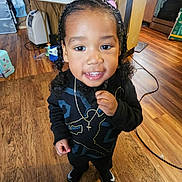 Tyreek is registered to the contest to win money with this photo: child, indoor, floor, wooden_floor, black_clothing, necklace, smile, curly_hair, braids, pigtails, person, young_child, happy, standing, home, flooring, toy, background_objects, household, casual_clothing