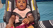 Sabrina is registered to the contest to win money with this photo: baby, child, car_seat, pink_headband, toy, striped_bedspread, blue_pillow, clothing, sitting, indoors, person, infant, cute, headband, seatbelt, bedroom, holding, footwear_absent, expression_neutral, young_child