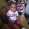 Sabrina is registered to the contest to win money with this photo: toddler, child, smile, pink_bow, shirt, love_text, pants, white_shoes, couch, leopard_print_pillow, blankets, indoor, happy, seated, person, curly_hair, home_interior, furniture, cozy, portrait