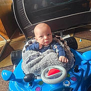 Xavier is registered to the contest to win money with this photo: baby, infant, child, face, portrait, sitting, walker, toy, blue, blanket, gray, clothing, indoors, living_room, furniture, carpet, steering_wheel, curious, baby_gear, photograph