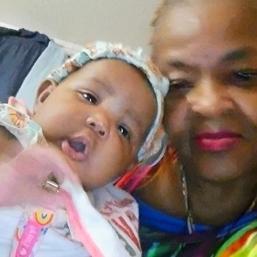 Mirah joined the competition — help win amazing prizes! baby, bonnet, casual, child, closeup, clothing, colorful, cross, face, family, floral_pattern, headwear, holding, indoor, necklace, person, portrait, skin, smile, woman