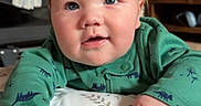 Jamie joined the competition — help win amazing prizes! baby, child, infant, green_clothing, dinosaur_pattern, face, hands, cushion, floral_pattern, indoor, wooden_furniture, cabinet, blanket, floor, curious_expression, bright_eyes, portrait, smile, soft_lighting, home