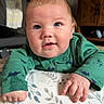 baby, child, infant, green_clothing, dinosaur_pattern, face, hands, cushion, floral_pattern, indoor, wooden_furniture, cabinet, blanket, floor, curious_expression, bright_eyes, portrait, smile, soft_lighting, home