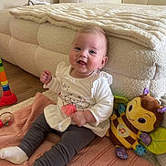 Ayla a rejoint le concours — aidez-le/la à gagner de superbes lots ! baby, child, smiling, sitting, blanket, toy, stuffed_animal, bee, couch, floor, white_clothing, gray_pants, happy, indoors, cute, person, infant, playful, soft_toy, home