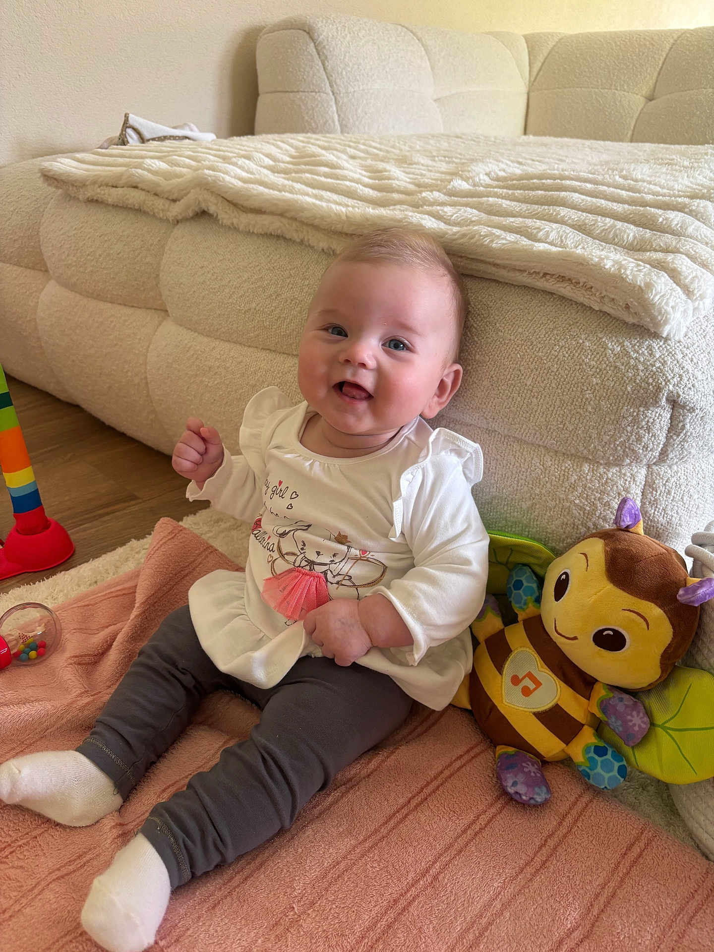 Ayla a rejoint le concours — aidez-le/la à gagner de superbes lots ! baby, child, smiling, sitting, blanket, toy, stuffed_animal, bee, couch, floor, white_clothing, gray_pants, happy, indoors, cute, person, infant, playful, soft_toy, home