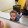 baby, mirror, reflection, medical_room, tummy_time, blue_clothing, person, phone, sink, cabinet, yellow_wall, white_wall, animal_decals, curly_hair, table, stool, hand, face, indoor, child