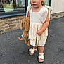 toddler, child, dress, stuffed_animal, toy, outdoor, pavement, window, brick_wall, sandals, curly_hair, expression, sidewalk, building, basket, footwear, person, summer, casual, young_child