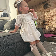Darcie-Rae joined the competition — help win amazing prizes! toddler, child, girl, white_dress, knee_high_socks, bow, shoes, couch, brick_wall, carpet, indoor, snack, cookie, hair_bow, furniture, toy, relaxed, side_view, casual, messy_floor