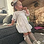 toddler, child, girl, white_dress, knee_high_socks, bow, shoes, couch, brick_wall, carpet, indoor, snack, cookie, hair_bow, furniture, toy, relaxed, side_view, casual, messy_floor