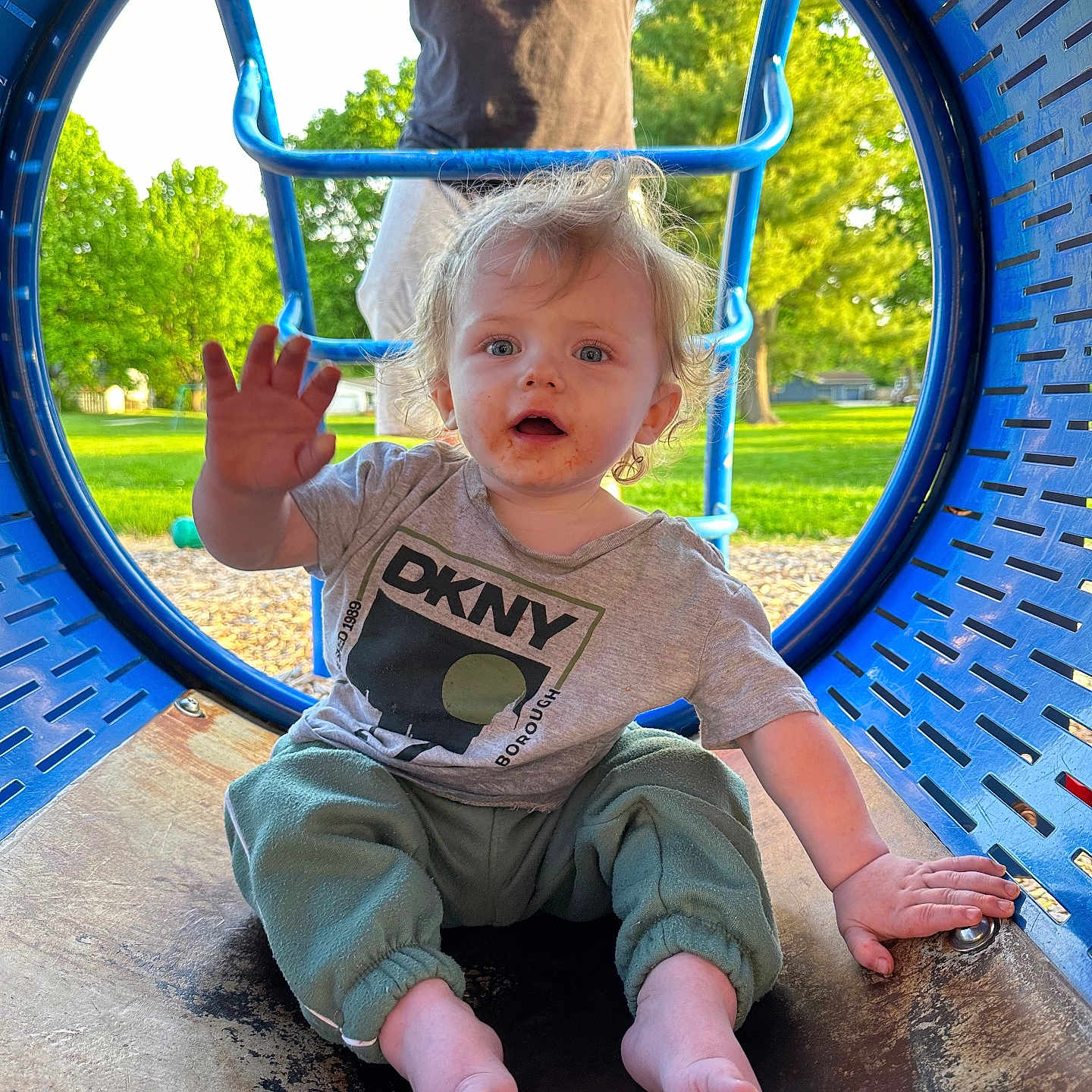 Stellar is registered to the contest to win money with this photo: toddler, child, playground, blue_tunnel, slide, greenery, park, outdoor, waving, messy_hair, casual_clothing, barefoot, person, daylight, happy, curious, sitting, adult, shoe, metal_surface