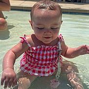 Gianna is registered to the contest to win money with this photo: baby, child, infant, swimming_pool, water, outdoor, summer, sunlight, pink_outfit, swimsuit, wet_hair, sitting, portrait, face, poolside, trees, lounge_chairs, reflection, shallow_water, happy