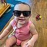 baby, child, sunglasses, smiling, sitting, pink_outfit, floor, wooden_floor, indoor, playroom, toy, toy_vehicle, portrait, face, person, infant, fashion, happy, closeup, daylight