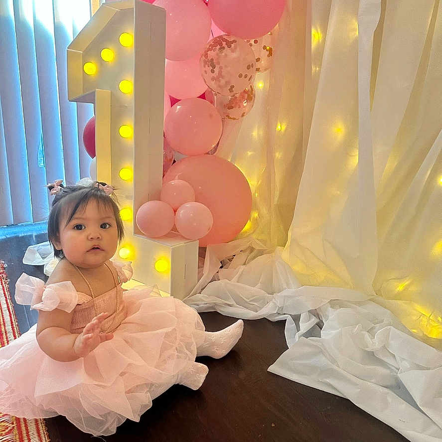 Sariyah joined the competition — help win amazing prizes! baby, balloons, birthday, celebration, child, curtains, cute, decorations, fairy_lights, festive, floor, girl, happy, indoor, number_one, party, pink_dress, portrait, sitting, toddler