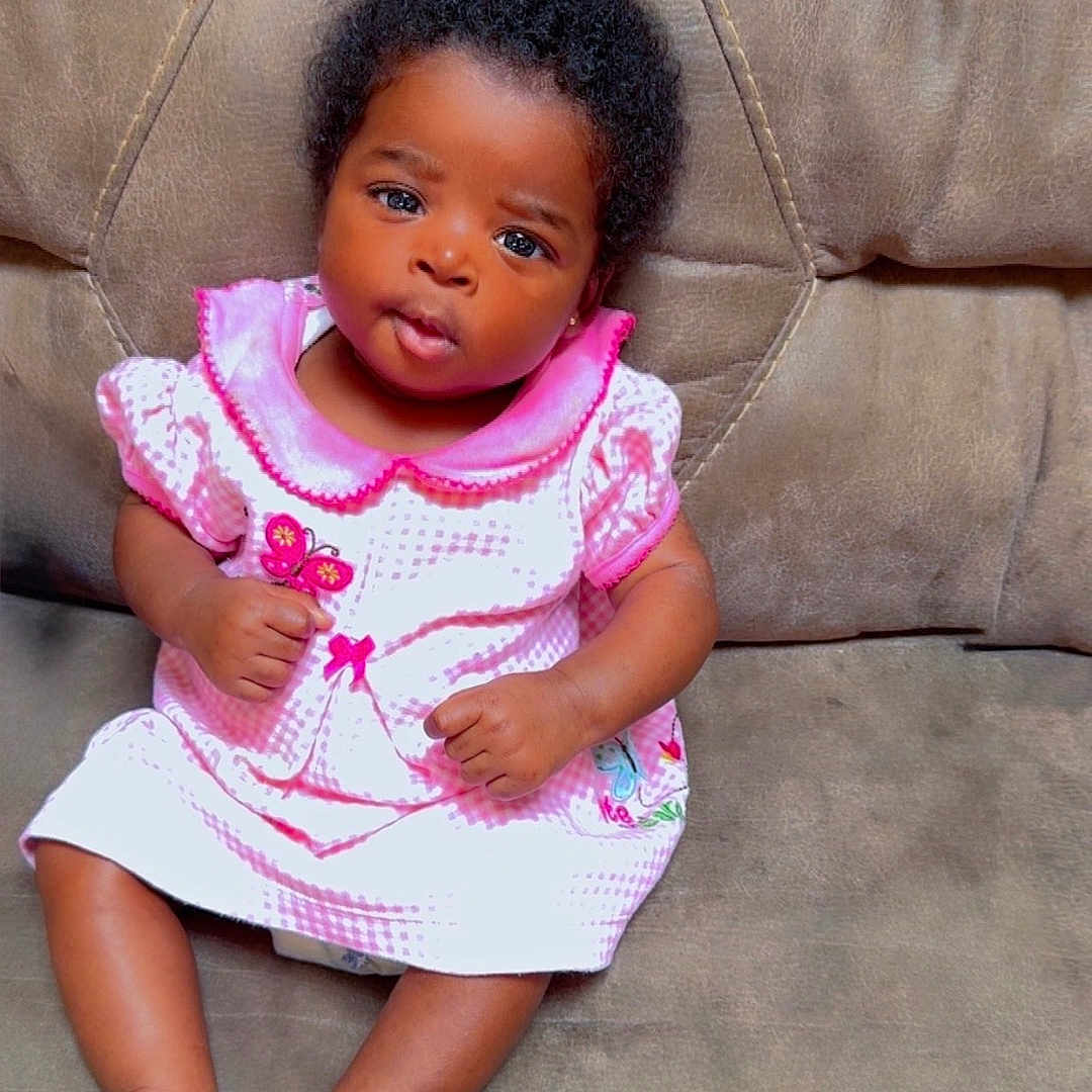 Jemima joined the competition — help win amazing prizes! baby, barefoot, butterfly, child, clothing, couch, curly_hair, cute, expression, face, furniture, indoors, infant, person, pink_dress, portrait, sitting, skin, sofa, young_child