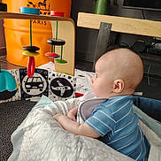 Maëlan C. a rejoint le concours — aidez-le/la à gagner de superbes lots ! baby, child, toy, wooden_toy, blue_clothing, striped_shirt, blanket, face, head, ear, potted_plant, orange_barrel, floor, living_room, television, table, curious, indoor, person, infant