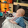 baby, child, toy, wooden_toy, blue_clothing, striped_shirt, blanket, face, head, ear, potted_plant, orange_barrel, floor, living_room, television, table, curious, indoor, person, infant