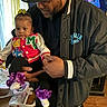baby, adult, indoor, holding, colorful_clothing, purple_bows, shoes, hat, jacket, television, smiling_faces, curtains, floor, wooden_floor, person, family, child, portrait, casual, home