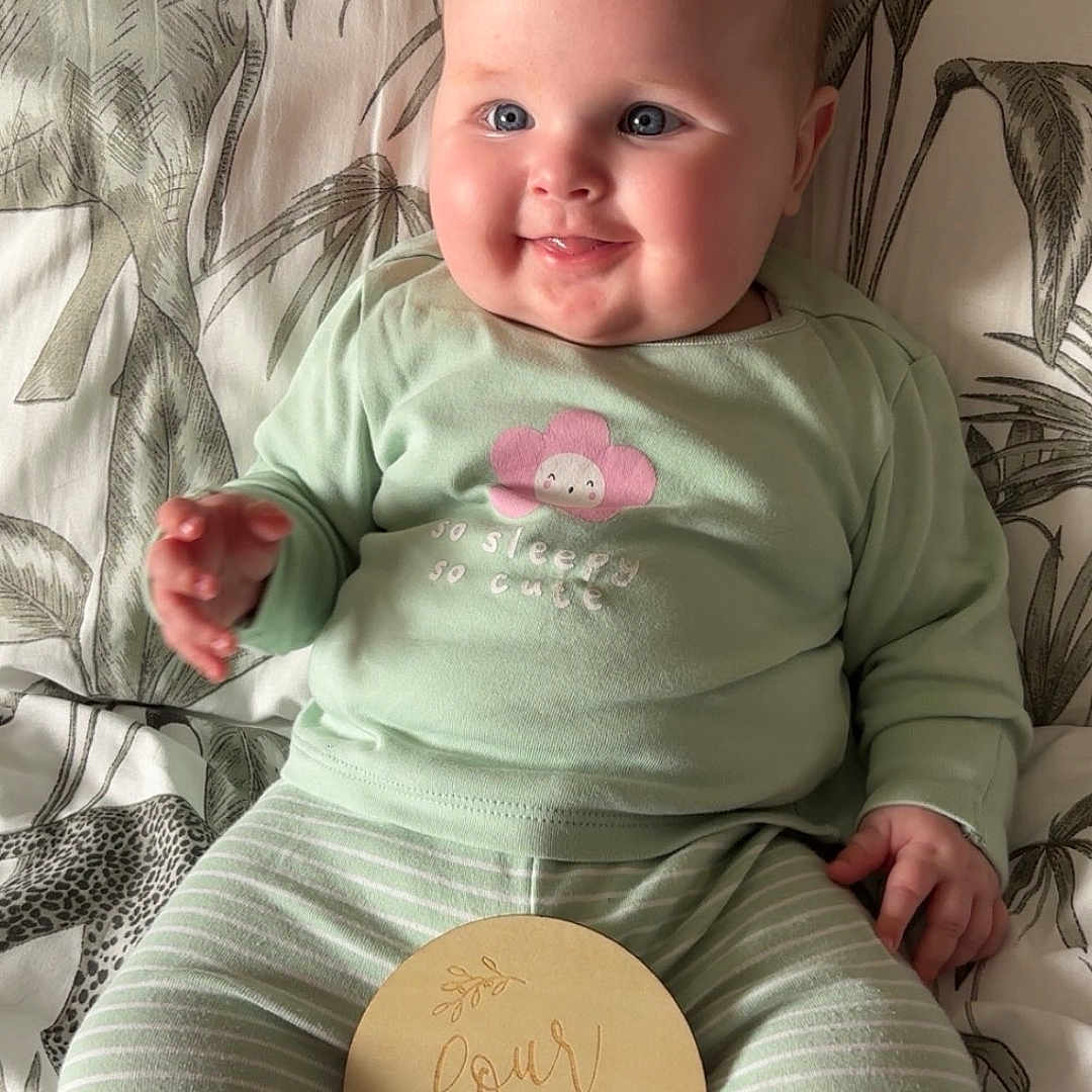 Florence is registered to the contest to win money with this photo: baby, bedspread, child, clothing, cute, face, feet, flower_design, four_months, hands, happy, indoor, infant, mint_green, patterned, portrait, sitting, smiling, striped_pants, wooden_sign