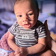 Kashmier is registered to the contest to win money with this photo: baby, infant, child, onesie, striped_clothing, text_on_clothing, face, eyes, mouth, hands, adult_hand, sitting, blanket, indoor, portrait, close_up, skin, expression, cute, bokeh