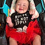 baby, car_seat, tutu, smiling, clothing, booties, infant, patterned, cute, happy, indoor, child, person, fabric, text_on_clothing, red, black, white, floral, seat