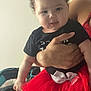 Zaria is registered to the contest to win money with this photo: baby, child, curly_hair, red_tutu, black_shirt, adult_hand, indoor, furniture, table, car_seat, person, holding, cute, smiling, portrait, infant, toddler, home, soft_lighting, clothing