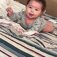 Zaria joined the competition — help win amazing prizes! baby, child, smiling, curly_hair, bed, blanket, striped_bedspread, indoor, person, happy, cute, lying_down, cozy, pillow, infant, wearing_clothes, face, hand, toddler, home