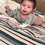 baby, child, smiling, curly_hair, bed, blanket, striped_bedspread, indoor, person, happy, cute, lying_down, cozy, pillow, infant, wearing_clothes, face, hand, toddler, home