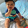 baby, child, infant, cap, toy, bed, quilt, lamp, indoor, smiling, chewing, colorful_clothing, shorts, sitting, hand_in_mouth, cute, happy, chubby, person, face
