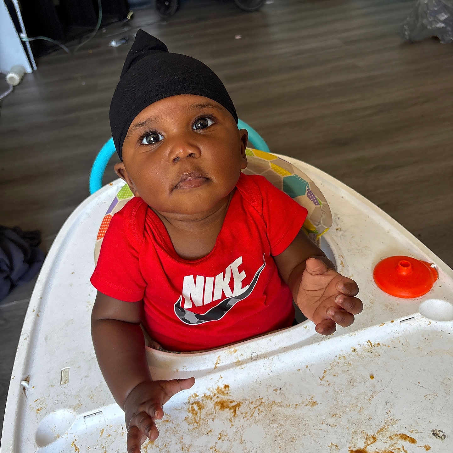 Tremeire joined the competition — help win amazing prizes! baby, child, high_chair, messy, food_stains, black_headwrap, red_shirt, nike_logo, indoor, flooring, curious_expression, hand_raised, face, person, young_child, seat, plastic_tray, toy_part, background_items, home_environment