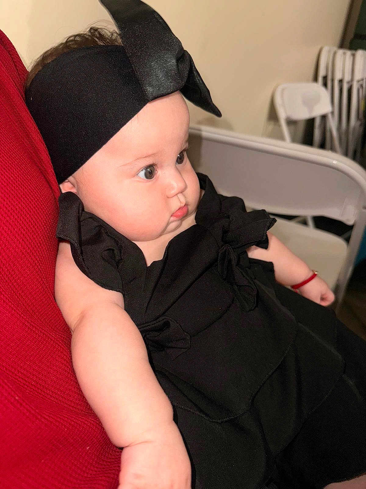 Luciana Vargas is registered to the contest to win money with this photo: baby, infant, black_clothing, headband, chubby_cheeks, curious_look, red_fabric, folding_chair, indoor, portrait, cute, child, person, sitting, arm, skin, young, headwear, closeup, fashion