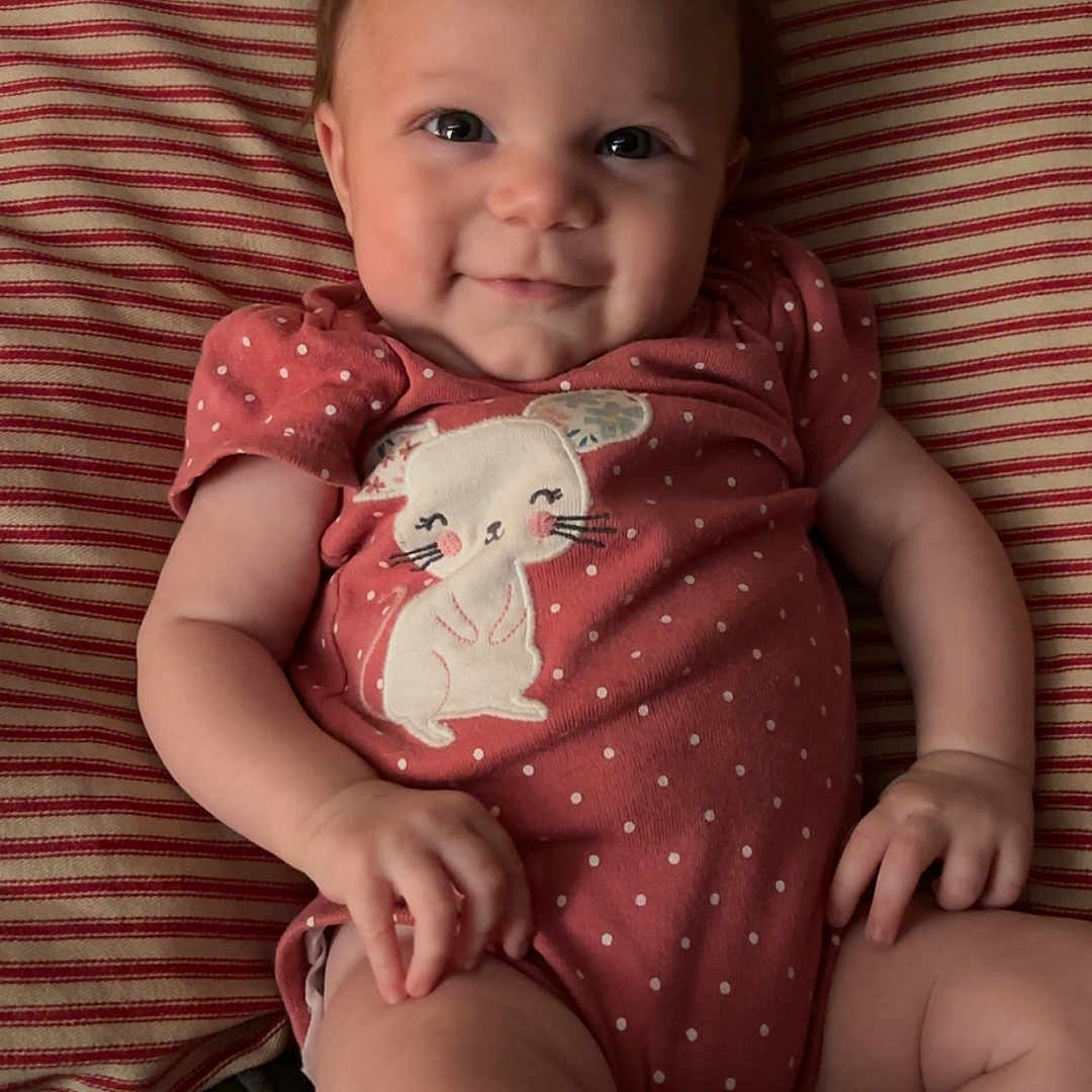 Holloni is registered to the contest to win money with this photo: baby, cat_design, cheeky, child, cute, face, hair_tuft, hand, happy, indoors, infant, lying_down, onesie, person, polka_dot, portrait, relaxed, skin, smile, striped_pillow