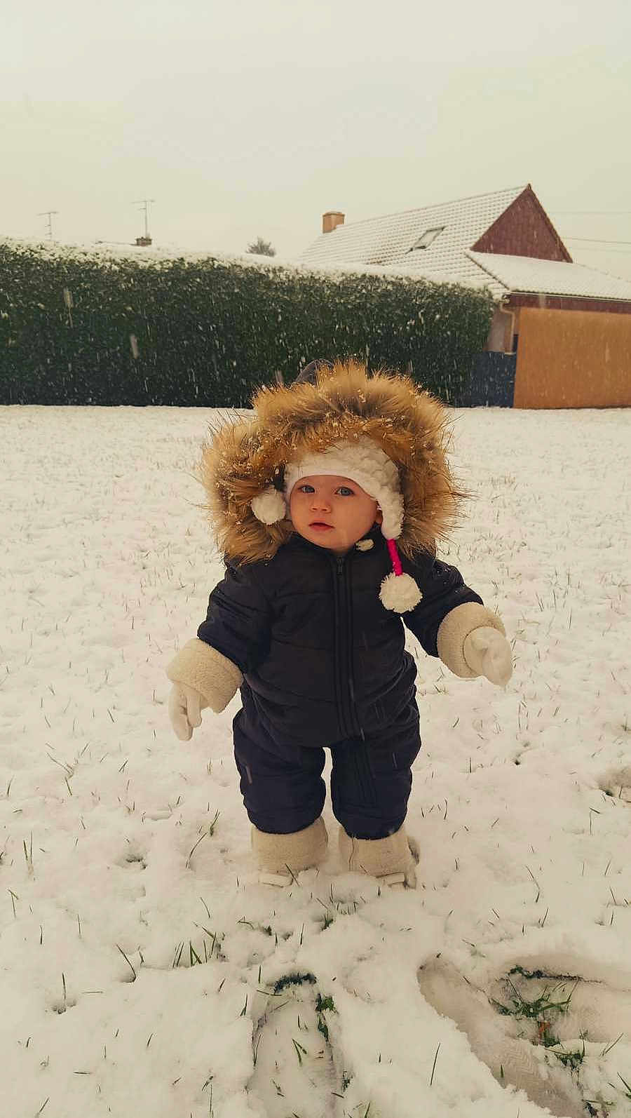 Léonnah participe au concours pour gagner de l'argent avec cette photo : baby, boots, child, cold_weather, curious, daylight, footprints, fur_hood, grass, hat, hedge, house, mittens, outdoor, portrait, snow, snowfall, snowy_ground, standing, winter_clothing