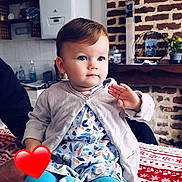 Léonnah participe au concours pour gagner de l'argent avec cette photo : child, toddler, baby, indoor, sitting, blue_leggings, patterned_dress, cardigan, hand_raised, face, bright_eyes, blanket, red_and_white, brick_wall, clock, furniture, plant, toy, person, cozy